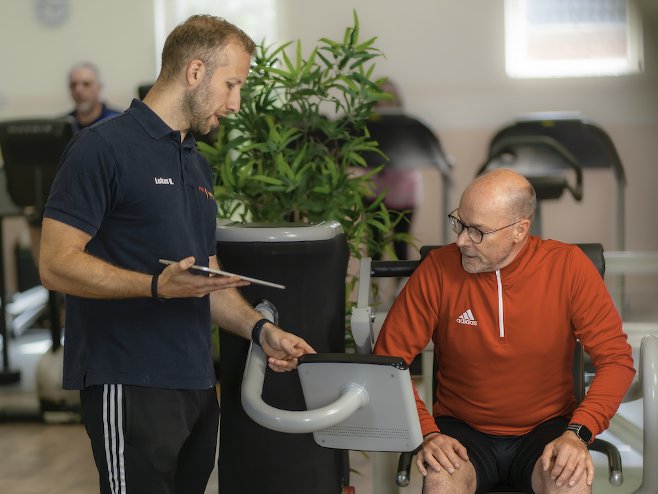 Lukas Heyer, Inhaber des Physioaktiv Sportsclubs, begleitet ein Mitglied beim Training am EGYM-Gerät. Er steht aufmerksam daneben, erklärt Funktionen und gibt motivierende Hinweise, während das Mitglied konzentriert die Übung ausführt. Im Hintergrund erkennt man die moderne, lichtdurchflutete Trainingsfläche des Studios.